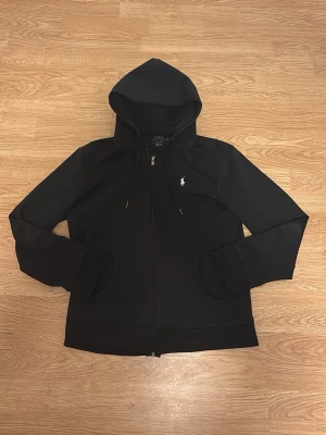 Svart hoodie från Polo Ralph Lauren  - Svart hoodie med dragkedja från Polo Ralph Lauren. Klassisk design med huva och snörning, vit broderad logga på bröstet och två fickor framtill. Tillverkad i mjuk bomull, perfekt för chill dagar. Långärmad och loose fit för skön komfort.