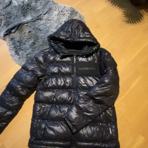 Svart pufferjacka Alessandro Zavetti - Snygg svart pufferjacka från Alessandro Zavetti med huva och dragkedja framtill. Jackan har en glansig finish och diskret logga på bröstet. Perfekt för kalla dagar när du vill hålla stilen och värmen.