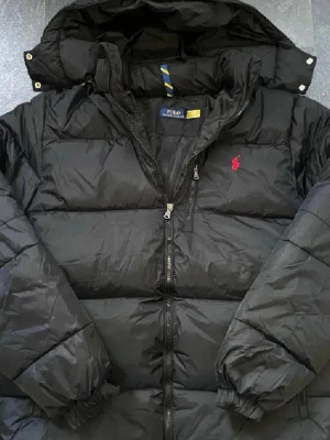 Svart pufferjacka från Polo Ralph Lauren - Svart pufferjacka från Polo Ralph Lauren med röd broderad logga på bröstet. Jackan har huva, RL-märkt dragkedja och breda quiltade sektioner. Perfekt för kalla dagar och har en modern, oversized vibe.