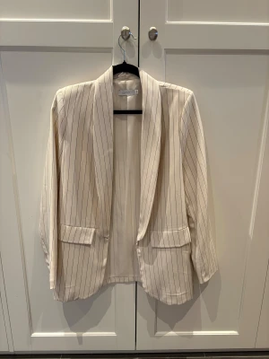 Randig beige kavaj från Sorbet - Snygg beige kavaj från Sorbet med tunna svarta ränder. Klassisk passform med två fickor framtill och lång ärm. Perfekt för dig som vill ha en stilren och trendig look. Tunn och lätt i materialet, passar året runt.