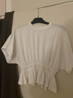 Vit croppad blus med volang - Säljer en vit blus med rund hals och korta ärmar. Toppen har en croppad passform med smockad midja och volang nedtill som ger en snygg siluett. Materialet är mjuk bomull och toppen är helt enfärgad utan mönster. Perfekt för dig som gillar stilrena och trendiga plagg.