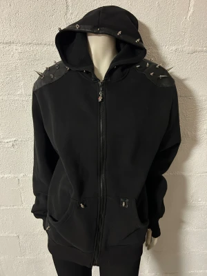 Huvjacka M fr Banned - Cool svart hoodie med dragkedja och huva. Den har nitar på axlar och ärmar för en edgy look. Dragkedjan har en dödskalle-detalj som ger extra attityd. Perfekt för dig som vill sticka ut! Storlek M. Märke Banned