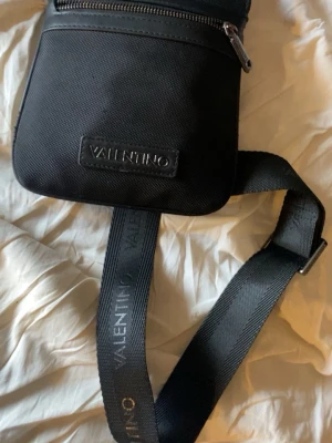 Svart axelväska från Valentino - Snygg svart axelväska från Valentino med justerbar rem där loggan är tryckt. Väskan har en stilren design med dragkedja upptill och är tillverkad i slitstark syntet. Perfekt storlek för det viktigaste och passar till en modern streetstil.