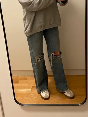 Blå slitna raka jeans från Zara - Säljer ett par blå jeans med raka ben och stora slitningar över knäna. Jeansen har hög midja, klassisk femficksmodell och råa kanter vid benslut. Perfekt vintage vibe och riktigt snygg passform.