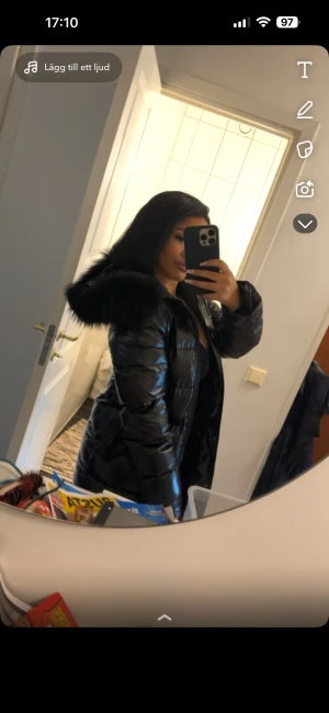 Svart lång pufferjacka med äkta päls - Säljer en svart, lång pufferjacka med glansig yta och stor huva med fluffig fuskpäls. Jackan har quiltad design, dragkedja framtill och praktiska fickor. Perfekt för kalla dagar och har en riktigt snygg streetstil. Passar dig som gillar oversized och mysiga vinterplagg. Obs har slitage som syns på bilderna