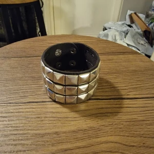 Svart skinnarmband med nitar - Säljer ett brett svart armband i skinn med tre rader silvriga pyramidnitar. 22 cm lång. Bredd 4,5 cm. Armbandet har tryckknappar för enkel stängning och ger en cool, edgy vibe till din outfit. Perfekt för dig som gillar rockig stil och vill sticka ut.