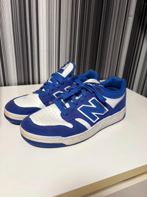 Blåvita New Balance 480 sneakers - Säljer ett par New Balance 480 sneakers i blått och vitt med klassisk N-logga på sidan. Skorna har snörning, perforerad tå och platt sula. Ovandelen är i en mix av mocka och syntet, med blå detaljer och vit bas. Perfekt för dig som gillar sportig stil. Storlek 43, lite använda men inga skador eller defekter eller sådant! 