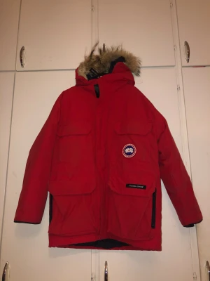 Röd Canada Goose parkas med päls - Riktigt fet röd parkas från Canada Goose med ikonisk patch på bröstet och stor huva med päls. Jackan har flera stora fickor, dragkedja och kardborre framtill. Perfekt för kalla vinterdagar när du vill hålla stilen och värmen. Ny pris 6000kr