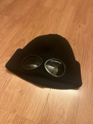 Svart mössa med goggles - Svart ribbstickad mössa med ikoniska goggles framtill. Snygg och urban design som sticker ut, perfekt för dig som gillar streetwear. Materialet är mjukt och värmande, och loggan syns tydligt på ena linsen. Grym accessoar för vintern!