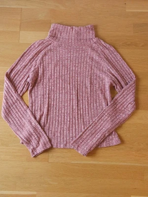 Rosa tröja - Säljer en mysig rosa tröja med ribbat mönster och hög krage. Perfekt för lager på lager. Materialet är mjukt och stretchigt, vilket gör den skön att ha på sig.