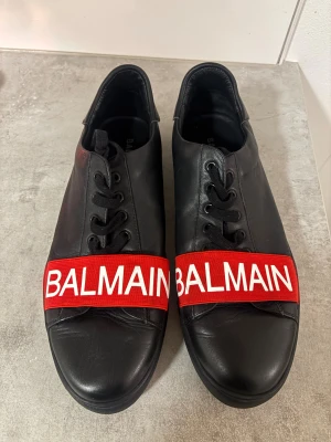 Balmain sneakers  - Säljer ett par svarta Balmain sneakers i skinn med feta röda band över tårna där det står BALMAIN i vitt. Skorna har svarta snören, rund tå och vit logga på hälen. Riktigt clean och lyxig vibe, perfekta för dig som vill sticka ut med designer på fötterna.