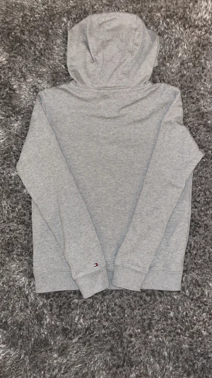 Grå hoodie Tommy Hilfiger - Snygg grå hoodie från Tommy Hilfiger med klassisk logga och text 'EST. 1985' på bröstet. Tröjan har huva, ribbade muddar och en stor magficka. Perfekt för chill dagar och enkel att matcha med jeans eller joggers. Pris kan diskuteras 