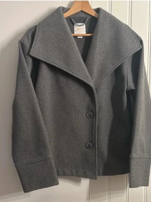Grå kappa med bred krage från Samsoe Samsoe - Säljer en stilren grå kappa från Samsoe Samsoe med bred krage och två stora knappar framtill. Jackan har lång ärm och är tillverkad i mjukt ullmaterial, perfekt för kyliga dagar. Enkel och snygg design som passar till många outfits.