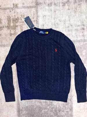Marinblå stickad tröja Polo Ralph Lauren - Snygg marinblå stickad tröja från Polo Ralph Lauren med klassiskt kabelstickat mönster och röd broderad logga på bröstet. Rund halsringning och långa ärmar. Perfekt för dig som gillar stilrena och tidlösa plagg.