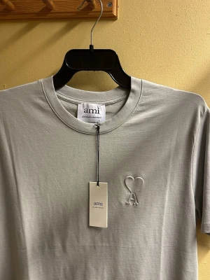 Ljusgrå t-shirt från Ami Paris - Snygg ljusgrå t-shirt från Ami Paris med broderad logga i form av ett hjärta och bokstaven A på bröstet. Klassisk rund halsringning och korta ärmar. Tillverkad i mjuk bomull för en skön känsla.