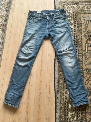 Replay Anbass jeans blå, storlek 33 - Snygga Replay Anbass jeans i blå tvätt med slitningar och hål på benet men är i fint skick annars. Modellen har klassisk femficksdesign, smal passform och raka ben. Jeansen är tillverkade i stretchigt denim för extra komfort och har detaljerade sömmar på bakfickorna.priset får att diskuteras.