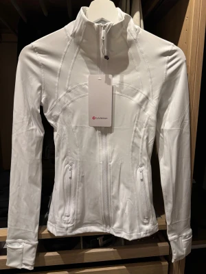 Vit träningsjacka från Lululemon storlek 2 - Supersnygg vit träningsjacka från Lululemon med figurnära passform, hög krage och dragkedja framtill. Jackan har två fickor med dragkedja och tumhål vid ärmsluten. Perfekt för gymmet eller powerwalks. Materialet är stretchigt och mjukt för maximal komfort. Fick den i present har inget kvitto därför säljer jag den lite billigare. Storlek 2 men passar en XS