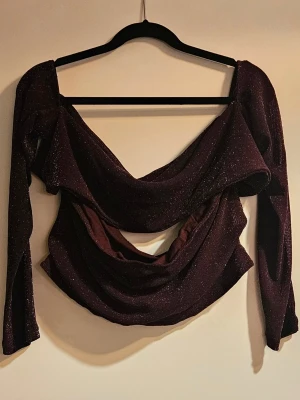 Glittrig mörklila topp - Snyggt glittrigt mörklila tvådelat set med en tubtop och en del med längre ärmar och knytdetalj framtill.. Setet har en croppad passform och är tillverkad i ett stretchigt material som skimrar i ljuset. Perfekt till nyår eller till utekvällen. Insåg i efterhand att jag fotat toppen fel och slänger in en produktbild från hemsidan som sista bild.