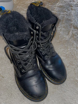  Dr. Martens kängor  - Säljer ett par svarta Dr. Martens boots med grov sula och gul söm runt kanten. Skorna har snörning och är fodrade med mjukt svart material för extra värme. Häl delen av skons material har lossnat off känns när man går i dom. Annars är skorna i fint skick 