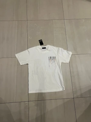 Vit AMIRI t-shirt med färgstänk - Säljer en vit t-shirt från AMIRI i mjuk bomull. T-shirten har korta ärmar och coolt tryck med AMIRI-logga och färgstänk i svart, orange, blå och turkos både fram och bak. Perfekt för dig som gillar streetwear och vill sticka ut med en unik design. Den är storlek Medium men passar som S eller XS. Den ska passa er som är runt 170-180.