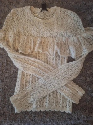 Crèmevit stickad tröja med volang - Supersöt vit pointellestickad tröja med volangdetalj över bröstet. Tröjan har ett genombrutet spetsmönster och långa ärmar. Perfekt för dig som gillar romantisk och feminin stil. Ribbad kant vid hals, ärmslut och nederkant.