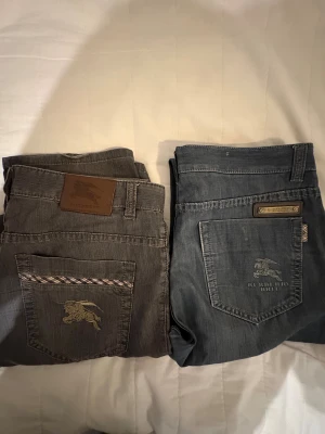 Burberry Brit jeans i grått och blått - Två par snygga Burberry jeans, ett i grått och ett i ljusblått. Båda har raka ben, klassisk femficksdesign och broderad Burberry-logga på bakfickan. Gråa paret har rutiga detaljer på fickorna och läderpatch i midjan. Perfekt för dig som gillar stilrena jeans med exklusiv känsla. Storlek W 34 L 32 på båda. 500 kr för ett par 850 kr för båda 