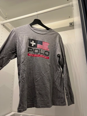 Grå långärmad topp från Polo Ralph Lauren - Snygg grå långärmad topp från Polo Ralph Lauren med tryck framtill i storlek L som inkluderar en amerikansk flagga och logga. Mjuk bomullskvalitet och rund hals. Perfekt för dig som gillar klassisk streetstyle med en sportig touch.