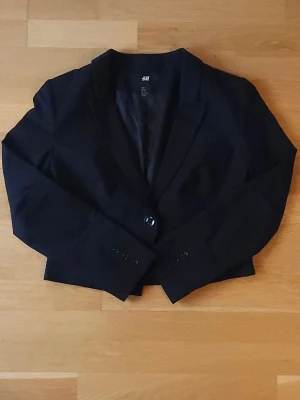 Svart kavaj - Svart, formsydd kavaj från H&M i croppad modell med klassisk krage och knapp framtill. Snygga detaljer med tre knappar vid ärmslutet och diskreta fickor. Materialet är mjukt och har en lätt glansig insida. Perfekt för dig som gillar stilrena och moderna plagg.