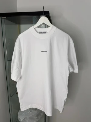 Vit Acne Studios t-shirt i bomull storlek M - Vit t-shirt från Acne Studios i 100% bomull med klassisk rund hals och korta ärmar. Minimalistisk design med liten svart Acne Studios-logga på bröstet. Perfekt för dig som gillar cleana och stilrena plagg.