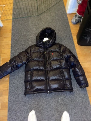 Svart dunjacka från Polo Ralph Lauren - Säljer en svart, glansig dunjacka från Polo Ralph Lauren med huva och två stora fickor med dragkedja. Jackan har en röd Polo-logga på bröstet och är perfekt för kalla dagar. Den är långärmad och har en riktigt schysst puffer-look.       Inköpt någon gång under förra vintern men bara använd ett fåtal gånger eftersom att den inte passar längre den är i nyskick och därav priset men även det går att diskuteras vid snabb affär såklart