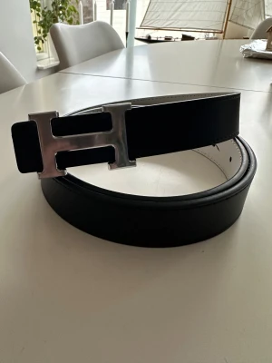 Svart och vit skinnbälte med H-spänne - Säljer ett vändbart bälte i svart och vitt skinn med ett stort, stilrent H-format metallspänne. Bältet har snygga sömmar längs kanterna och är 120 cm långt. 