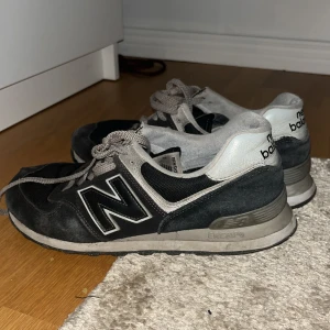 Svarta New Balance 574 sneakers - Säljer ett par klassiska New Balance 574 sneakers i svart och grått med stor N-logga på sidan. Skorna har snörning, platt sula och rund tå. Ovandelen är i mocka och mesh, medan hälen är i syntetmaterial. Perfekta för dig som gillar retrostil och streetwear.