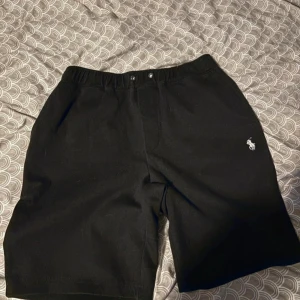 Svarta shorts från Polo Ralph Lauren - Svarta shorts från Polo Ralph Lauren med klassisk logga broderad i vitt på ena benet. Modellen har elastisk midja och två sidofickor. Materialet är mjuk bomull som känns skönt mot huden. Perfekta för chill dagar eller när du vill ha en clean look.