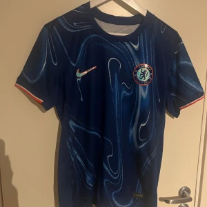 Chelsea 24/25 - Chelsea FC matchtröja från Nike i marinblått med unikt abstrakt mönster. Kortärmad modell med rund hals och klubbmärke på bröstet. Materialet är lätt och andas, perfekt för träning eller match. Orange och vit detalj på ärmslut och Nike-logga på framsidan.
