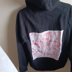 Svart hoodie med tryck och huva - Svart hoodie med stor huva och snörning. På ryggen finns ett stort vitt tryck med röda läppavtryck. Tröjan har en klassisk känguruficka framtill och långa ärmar. Perfekt för dig som gillar streetstyle och unika detaljer.