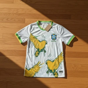 Vit Brasilien fotbollströja Nike - Vit fotbollströja från Nike med Brasilien-emblem på bröstet och stora gula fåglar tryckta framtill. Gröna och gula detaljer på krage och ärmslut. Tillverkad i lätt och ventilerande material, perfekt för match eller träning.