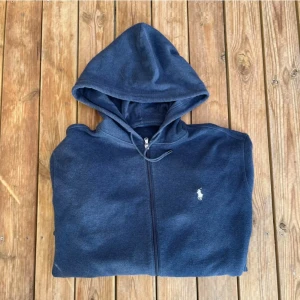 Ralph Lauren Hoodie - Säljer en mörkblå hoodie från Ralph Lauren med dragkedja och huva. Tröjan har två fickor framtill med dragkedja och den klassiska loggan broderad på bröstet. Perfekt för en avslappnad stil.