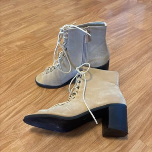Beige mocka boots med klack och snörning - Snygga boots i beige mocka med kraftig klack och snörning framtill. Skorna har rund tå, dragkedja på sidan och platt sula. Perfekt för dig som gillar en cool och trendig look med lite höjd. Passar till både jeans och kjol.
