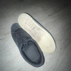 Svarta sneakers från Selected Homme - Säljer ett par svarta sneakers från Selected Homme i mocka med vita sulor och klassisk snörning. Skorna har en stilren och enkel design med rund tå och platt sula, perfekta för en clean look. Storlek 42.