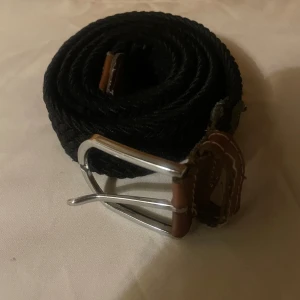 Svart flätat bälte med skinn och metallspänne - Svart flätat bälte med detaljer i brunt skinn och ett stilrent silverfärgat metallspänne. Bältet har vita sömmar på skinnpartierna och en klassisk rektangulär form på spännet. Perfekt för att ge outfiten en snygg touch.