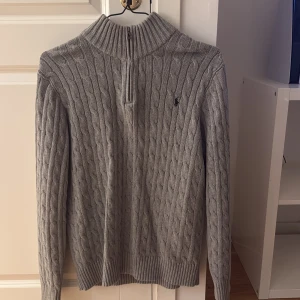 Grå stickad tröja Polo Ralph Lauren - Kabelstickad grå tröja från Polo Ralph Lauren med half zip och hög krage. Klassisk design med broderad logga på bröstet och ribbade muddar. Perfekt för lager-på-lager och chill höst- och vinterstil.