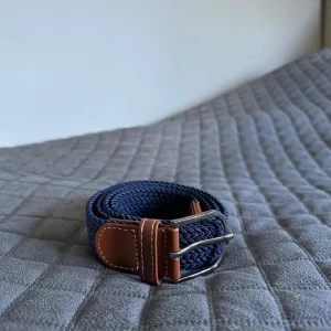 Mörkblå flätat bälte med skinn 105cm - Snyggt mörkblått flätat bälte med detaljer i brunt skinn och silverfärgat spänne. Bältet har en stilren look med kontrastsömmar på skinnet och passar perfekt till jeans eller chinos. Flätad design gör det enkelt att justera längden efter behov. 105 cm