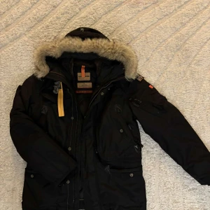 Svart Parajumpers vinterjacka med päls - Grym svart vinterjacka från Parajumpers Masterpiece Series med pälsfodrad huva, flera fickor och snygga detaljer som gul rem och patch på ärmen. Jackan har dragkedja, knappar och är perfekt för kalla dagar. Slim fit-modell med robust känsla.