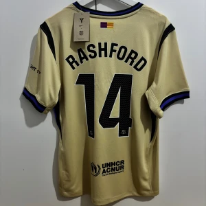  FC Barcelona borta 25/26 # Rashford 14 Player Edition  - Säljer en FC Barcelona fotbollströja med Rashford och nummer 14 på ryggen. Tröjan har korta ärmar, svarta och blå detaljer, samt tryck från Spotify och UNHCR. Materialet är lätt och andas, perfekt för match eller träning. Den är Player Edition en av de högsta kvalitet TVEKA INTE ATT SKICKA EN BUD