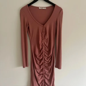 Rosa/brun ribbad långärmad klänning NA-KD - Supersnygg långärmad klänning från NA-KD i en dämpad rosa/brun nyans. Klänningen har v-ringning, ribbat material och dekorativa knappar med rynk framtill. Figurnära passform och stretchigt tyg som sitter bekvämt. Perfekt för dig som gillar stilrena detaljer.