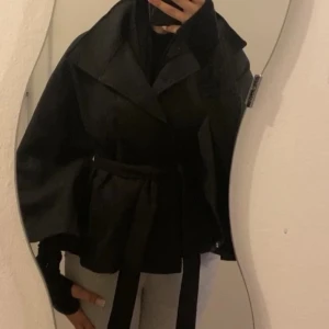 Svart poncho jacka med bälte! - Snygg svart poncho jacka med bred krage och bälte i midjan som ger en cool siluett. Jackan har en loose passform och långa ärmar, perfekt för dig som gillar en stilren och enkel look. Materialet känns mjukt och har en matt finish.