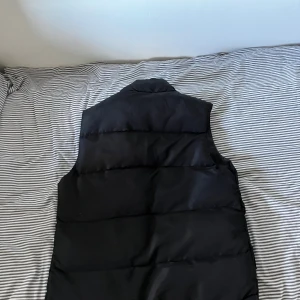 Svart dunväst från Canada Goose - Svart dunväst från Canada Goose med klassisk logga på bröstet. Västen har hög krage, stora fickor framtill och stängs med både dragkedja och knappar. 