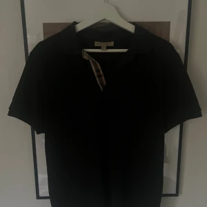 Burberry Piké - Snygg svart piké från Burberry med klassisk krage och knappslå i det ikoniska rutiga Burberry-mönstret. Storlek M, sitter som en M.