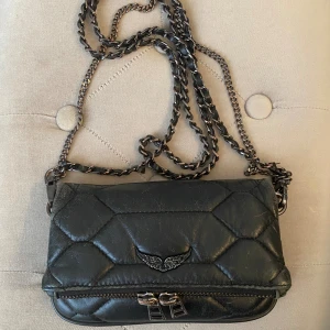 Svart quiltad axelväska Zadig & Voltaire - Snygg svart quiltad axelväska från Zadig & Voltaire i skinn med cool kedjerem och metall-detaljer. Väskan har ett emblem med vingar på framsidan, dragkedja både fram och bak samt en kompakt form som passar perfekt för det viktigaste. Kedjan har inslag av läder för extra edge.