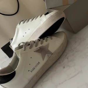 Golden Goose vita sneakers stl 43 - Golden Goose sneakers i vitt skinn med grå mockadetaljer och grå snörning. Klassisk stjärna på sidan, svart hälparti med logga och platt sula. Snygg och stilren modell som passar till det mesta. (Passade inte mig)